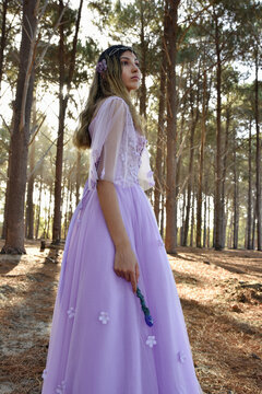 portrait of beautiful young blonde model wearing a purple princess fantasy ball gown with flower crown diadem.  Holding magical crystal wan din pine tree forest location background with golden lightin