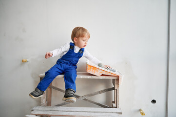 Cute kid construction worker sitting on wooden table against white wall in apartment under renovation. Cheerful little boy wearing safety helmet and work overalls while playing at home.