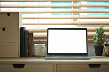 Front view laptop, books, coffee cup and houseplant on wooden counter with sunlight from blinds window