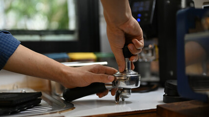Professional male barista holding portafiler, using coffee tamper to press coffee powder