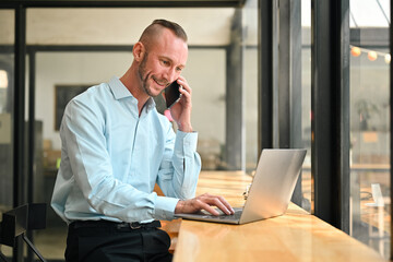 Obraz premium Smiling caucasian businessman talking on mobile phone and checking information on laptop computer at coffee shop