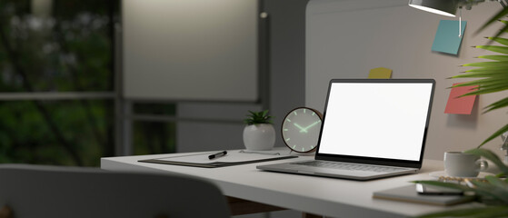 Close-up image of modern office desk with laptop mockup, stationery, clock, table lamp, and decor.