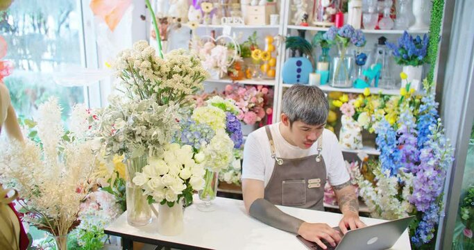 Asian male florist shop owner looking and typing on laptop making order. Tattooed guy hipster seller in apron sitting in floral store working on computer. SME Small business man entrepreneur.
