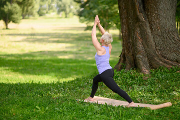 Middle aged caucasian woman in sportswear doing yoga outdoors in warrior pose, Virabhadrasana II. The concept of yoga, stretching, healthy lifestyle. Sports activities in nature. Copy space. 