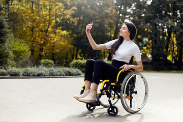 Obraz premium Attractive young woman sitting in a wheelchair with a phone in her hand. Beautiful girl smiling while taking a selfie on the phone outdoors.