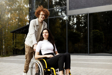 Cheerful red-haired, curly guy in glasses and braces carries a wheelchair with a girl. A beautiful brunette sits and looks away, on the street.