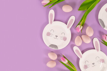 Easter party flat lay with bunny shaped paper plates, eggs and tulip flowers on violet background with copy space