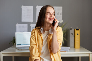 Photo of dreaming smiling woman with brown hair, fem ale wearing casual clothes and has headphones,...