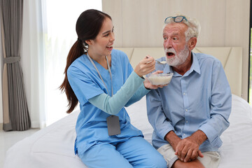 Happy caregiver feeding senior old man on bed with rice porridge soup for breakfast at retirement...