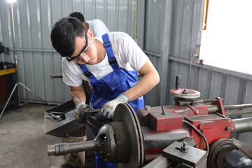 Multiethnic technicain mechanic or worker man in protective uniform using metal lathe machine operate polishing car disc brake at garage. Maintenance automotive and inspecting vehicle part concept