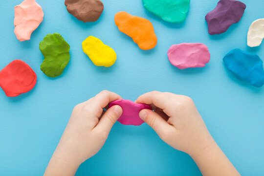 Baby boy hands holding and kneading modeling clay on light blue table background. Pastel color. Closeup. Top down view. Toddler development. Preparing material for making different colorful shapes.