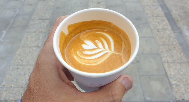 Holding A Delicious, Golden Brown Flat White Coffee With Foamy Flower Coffee Art In Takeaway Cup