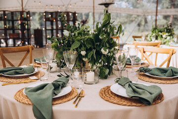 Wedding. Banquet. On the festive table with a tablecloth is a composition of flowers and greenery, there are plates with green napkins on wicker stands, glasses, cutlery and candles in glass vases