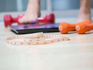 Closeup of measuring tape with blurry orange, red dumbbells,  weight scale on wooden floor and women 's foot for dieting or healthy concept