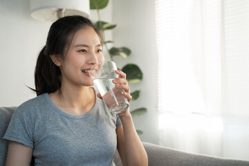 Healthy beautiful Asian woman is drinking and sip glass of fresh water everyday routine for body hydration by holding transparent glass in her hand, feel thirsty at home in the morning