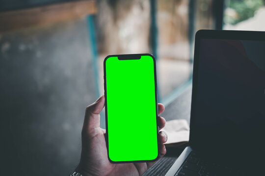 Mockup image of a business man holding smart mobile phone with blank green screen on vintage wooden table in modern cafe restaurant during meeting or lunch..
