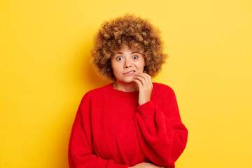 Fototapeta na wymiar Worried embarrassed woman with Oops expression looks scared and guilty bites lips keeps fingers on chin looks nervously at camera dressed in red pullover isolated against yellow background