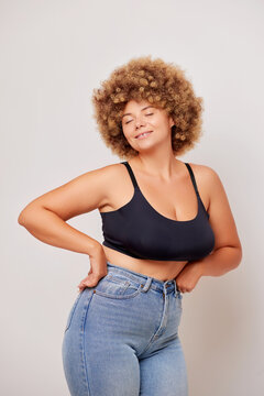 Sideways shot of self joyed sensual body positive woman with curly hair holds arms on hips feels confident in own body closes eyes and smiles gently wears basic lingerie bra and blue jeans poses over