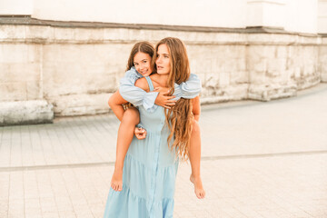 Mother of the daughter walks playing. Mother holds the girl on her back, holding her legs, and her daughter hugs her by the shoulders. Dressed in blue dresses.