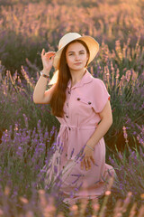 A cheerful young girl in a straw hat and a pink dress sits and rests among lavender bushes. Sunset.