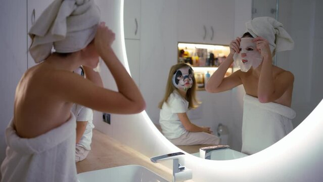 Mother applying tissue panda mask on her face relaxing in the bedroom with her daughter next to her