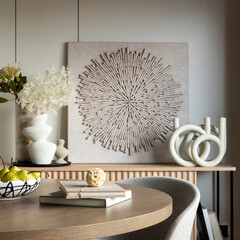 Interior design of aesthetic and elegant room with stylish commode, round table, grey chair, flowers in modern vases, beautiful paintings, candlestick and elegant personal accessories. Home decor.