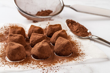 Chocolate truffles sprinkled with cocoa powder closeup. Delicious dark chocolate candy balls, mesh strainer and spoon over marble surface. Homemade sweet food dessert recipe concept.