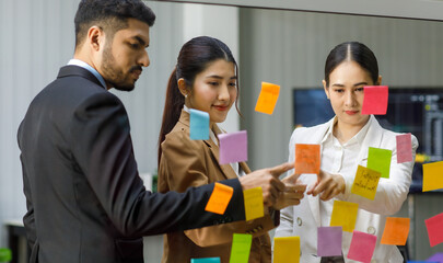 Millennial Asian Indian professional successful male businessman and female businesswomen colleagues in formal business suit standing pointing post it sticky note on glass board discussing together