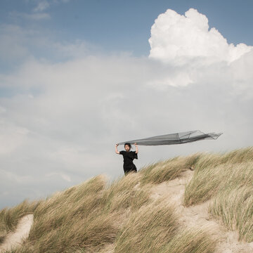 girl in black dress standing on dune holding fabric blowing in the wind and clouds