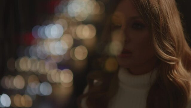 View of tender beautiful girl watches lights of night city at window in her bedroom, apartment. Car lights reflected in the glass, bokeh. Missing of somebody loved, waiting and worrying. HQ 4k footage
