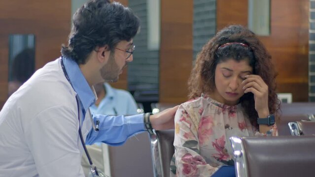 A helpful and supportive Indian Asian male doctor wearing an apron and stethoscope checking file, talking and consoling a sad and worried female relative of patient sitting in a hospital waiting area