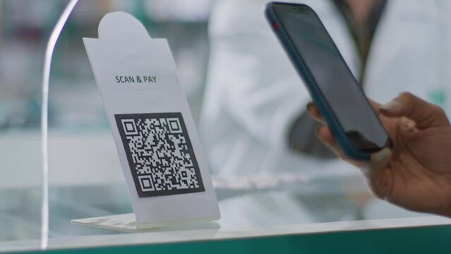 A close-up shot of a hand of a customer scanning a barcode to pay e-money to an electronic bank account or online UPI transfer in a general or grocery store with a shopkeeper in the background.
