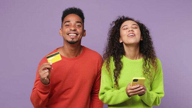 Young couple two friends family man woman of African American ethnicity wear casual clothes together use mobile cell phone hold credit bank card do online shopping isolated on plain purple background