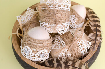 Easter eggs wooden, decorated with lace and braid close-up in a wooden plate on a light yellow background. Handmade rustic style