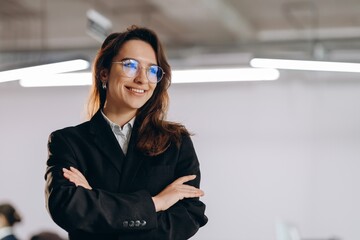 Portrait of smiling manager girl wearing glasses and black suit. Happy businesswoman standing with arms folded and looking away