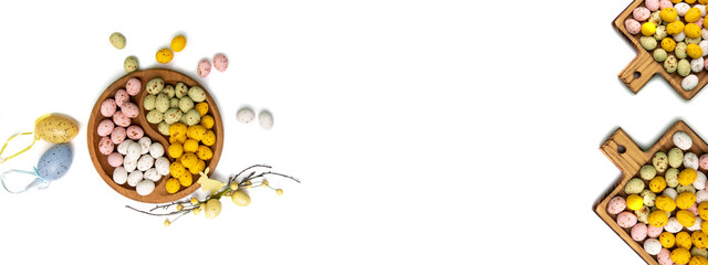 Multi-colored chocolate decorative eggs in wooden plate on a white background with decorative branches. Happy Easter background, web banner. Flat lay