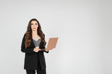 Curly adult business lady in elegant jacket standing while working on laptop against gray background