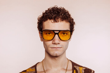 Closeup portrait of young stylish homosexual man with curly hair, wears orange sunglasses at studio isolated over beige background. Fashion concept.