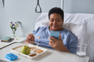 Portrait of black senior woman eating healthy meal in hospital room and using phone
