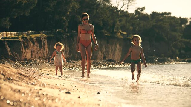 Mom and her little kids walk along the sea beach together, slow motion. Summer vacation time