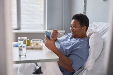 Side view portrait of smiling senior woman using phone in hospital room and eating breakfast