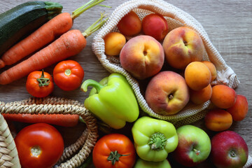 Straw bag and reusable fabric bags filled with various healthy fruit and vegetables. Wooden background, top view.