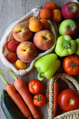 Straw bag and reusable fabric bags filled with various healthy fruit and vegetables. Wooden background, top view.