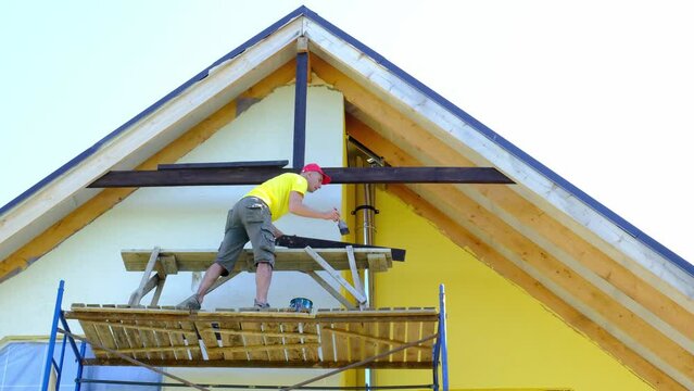 Male builder makes exterior decoration of private house in the style of half-timbered house with his own hands - he mounts wooden beams on facade, pediment under the roof high on the scaffolding