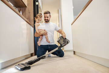 A tidy father is vacuuming floor in a kitchen while babysitting his baby girl.