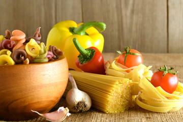 Italian food cooking ingredients. Pasta, vegetables, spices. Uncooked spaghetti, noodles, bell pepper, tomato, garlic, macaroni in wooden mortar on wooden background. Culinary concept. Close-up view