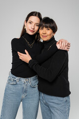 asian mother and daughter in black turtlenecks and jeans embracing and looking at camera isolated on grey