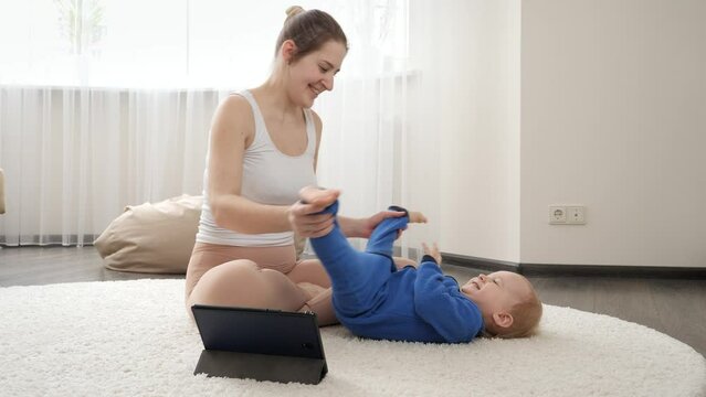 Young Mother Dwatching Online Video Course And Stretching Her Baby Boy Body On Carpet At Home. Family Healthcare, Active Lifestyle, Parenting And Child Development.