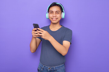 Young Asian man in a casual t-shirt wearing headphones for listening to music and holding mobile phone isolated over purple background