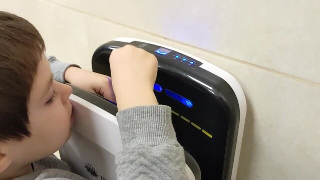A 5 year old boy dries his hands in an electric hand dryer in public bathroom.